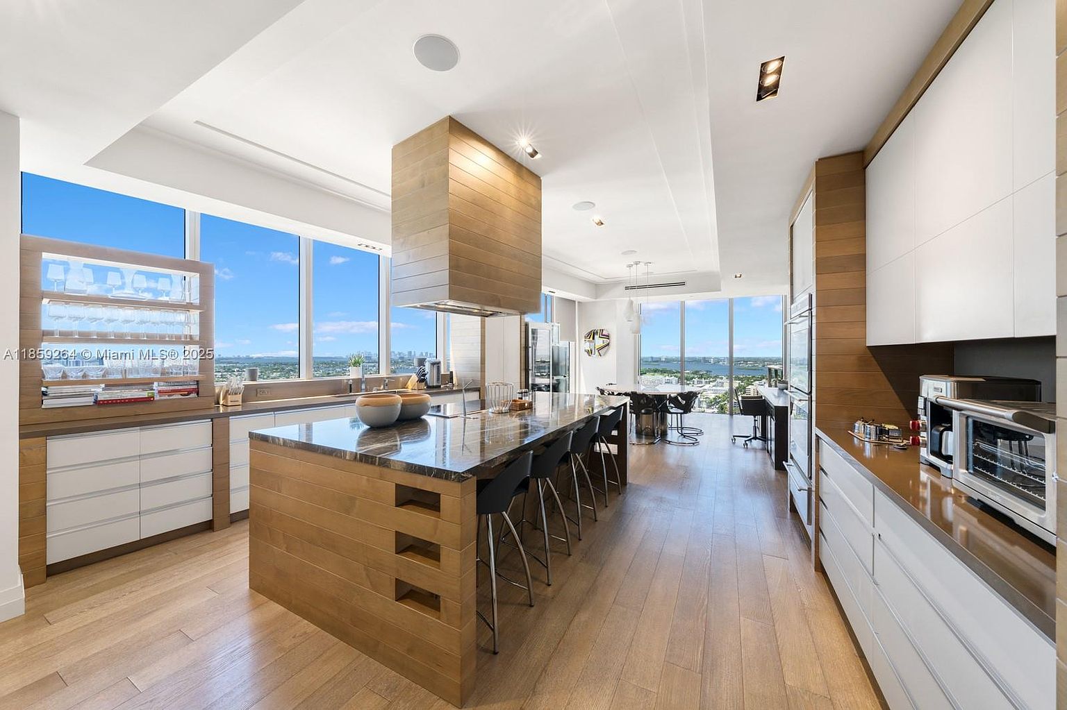 This is a bright and modern kitchen featuring a large island with seating, wood-paneled accents, and white cabinetry. The kitchen boasts floor-to-ceiling windows offering expansive views, and the open layout connects seamlessly to the dining area. The overall impression is one of luxury and sophistication.