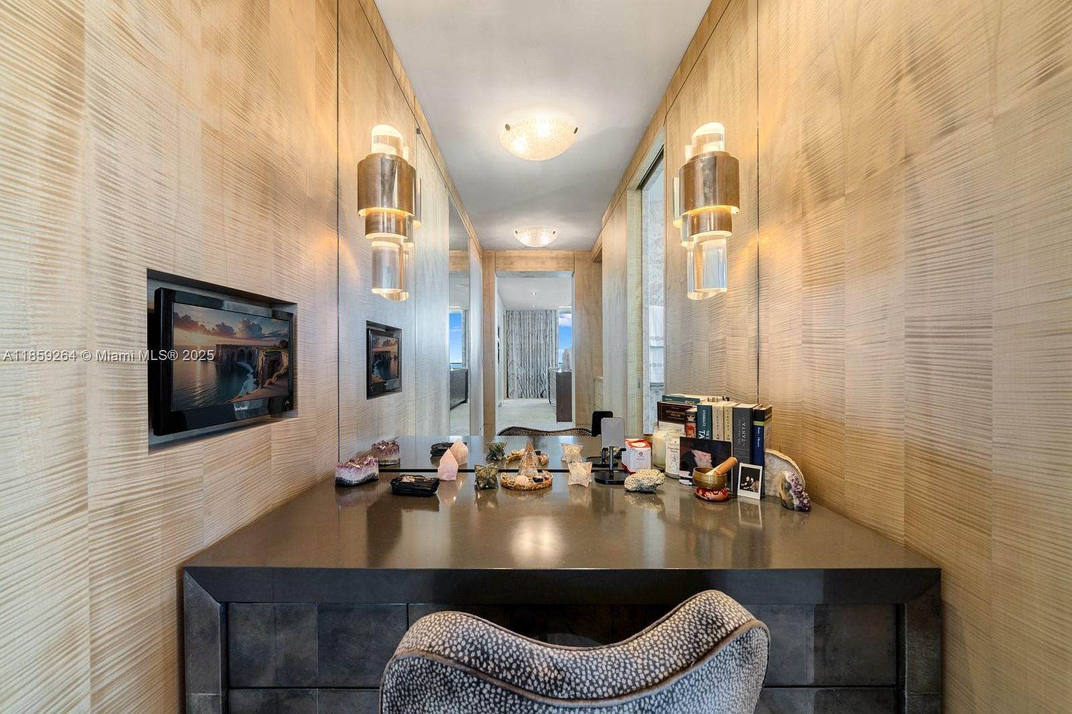 This interior shot showcases a stylish hallway or dressing area with mirrored walls, creating an illusion of depth and space. A dark-toned desk or vanity is adorned with decorative items, and modern sconces provide warm lighting. The textured wallpaper and unique chair add to the room's sophisticated and contemporary aesthetic.