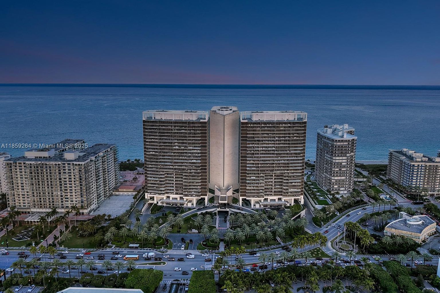 This aerial view showcases a luxury beachfront property featuring multiple high-rise buildings with a modern architectural style. The buildings are surrounded by lush landscaping, including palm trees and well-manicured lawns, with roads and traffic visible below. The ocean provides a stunning backdrop, creating an impression of exclusivity and upscale living.