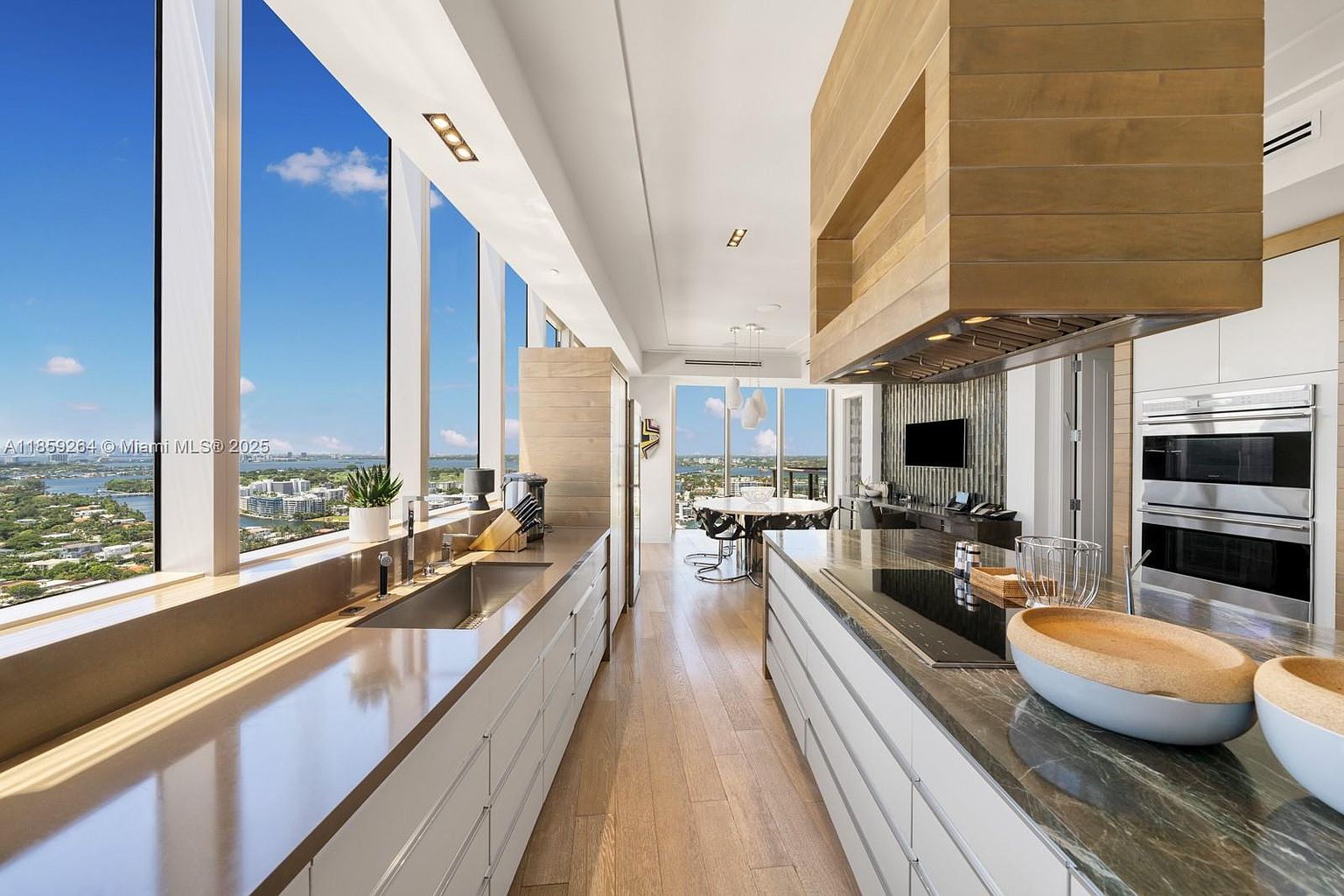 This is a bright, modern kitchen with floor-to-ceiling windows offering a stunning city view. The kitchen features sleek white cabinetry, stainless steel appliances, and a large island with a dark countertop. The open layout connects to a dining area, creating a spacious and inviting atmosphere.