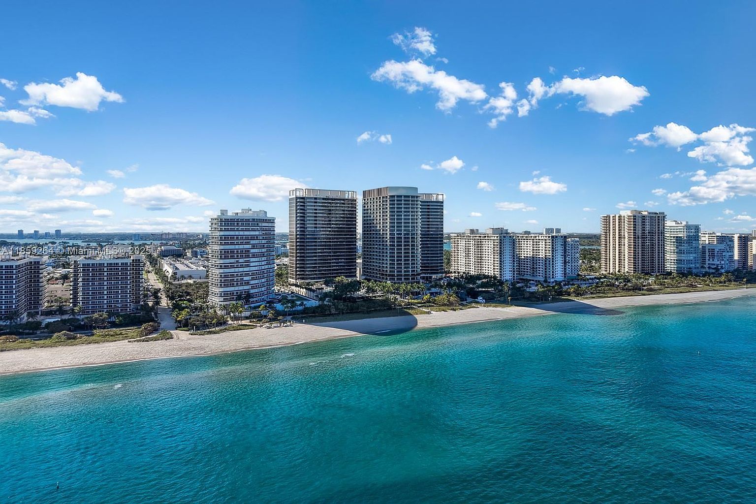 This aerial view showcases a stunning beachfront property featuring multiple high-rise condominium buildings along a pristine sandy beach. The turquoise ocean water contrasts beautifully with the clear blue sky dotted with fluffy white clouds. The image emphasizes the desirable coastal location and luxurious living environment.