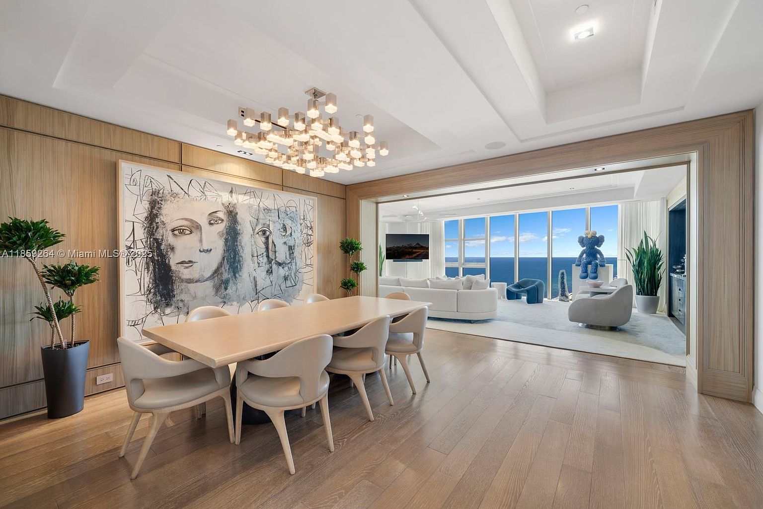 This interior shot showcases a luxurious dining room that seamlessly transitions into a living area with ocean views. The dining room features a large, light wood table surrounded by modern chairs, illuminated by a striking contemporary chandelier. A large abstract artwork adorns the wall, adding an artistic touch to the space, while the open layout leads to a bright living area with floor-to-ceiling windows overlooking the ocean.