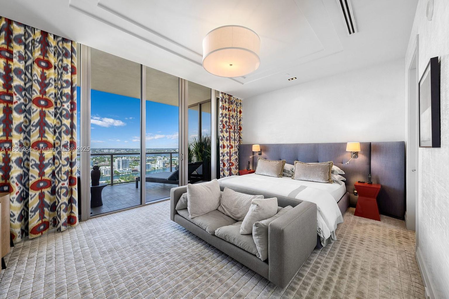 This is a primary bedroom featuring a large bed with a gray headboard, flanked by red side tables and lamps. A gray sofa sits at the foot of the bed, and patterned curtains frame the sliding glass doors leading to a balcony with city views. The room is well-lit with a modern ceiling light fixture and has a luxurious, contemporary feel.