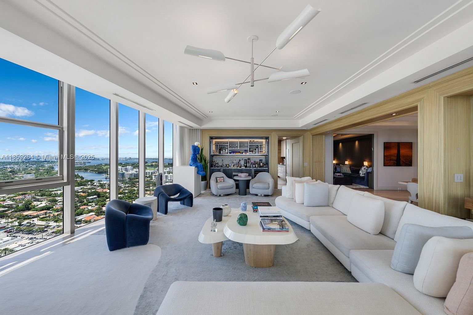 This is a luxurious living room featuring floor-to-ceiling windows with a stunning city view. The room is furnished with a large white sectional sofa, modern armchairs, and a unique coffee table. A built-in bar area adds a touch of sophistication, and the overall design is contemporary and elegant.