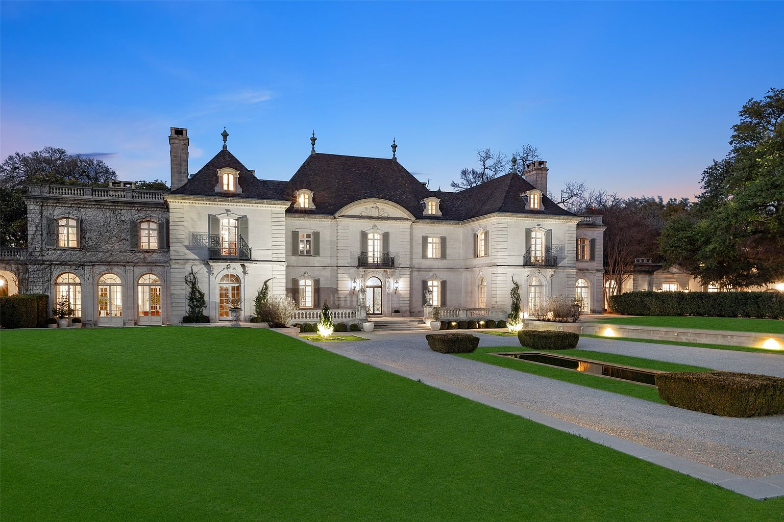 This is a stunning front view of a grand estate, showcasing its elegant architecture and meticulously landscaped grounds. The building features a symmetrical facade with multiple windows, balconies, and a dark roof, all illuminated by soft lighting. A sprawling green lawn leads to a gravel driveway with manicured hedges and water features, creating a luxurious and inviting atmosphere.