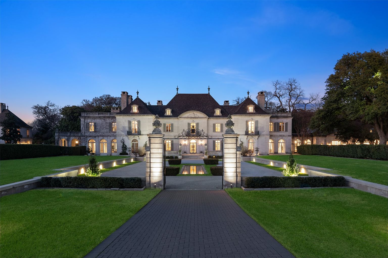 This is a stunning front view of a grand estate at dusk. The architecture is reminiscent of a French chateau, featuring a symmetrical facade, multiple dormers, and a dark roof. The property is meticulously landscaped with manicured lawns, hedges, and a formal entrance with decorative gates and lighting, creating an impressive and inviting approach.