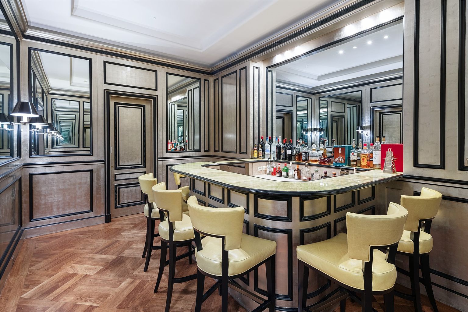 This is an interior shot of a sophisticated wine cellar featuring a bar area with several bottles of liquor displayed on the counter. The walls are decorated with mirrored panels and dark trim, creating an illusion of depth and luxury. The bar is surrounded by stylish chairs, and the floor is a herringbone pattern wood, adding to the room's elegant ambiance.