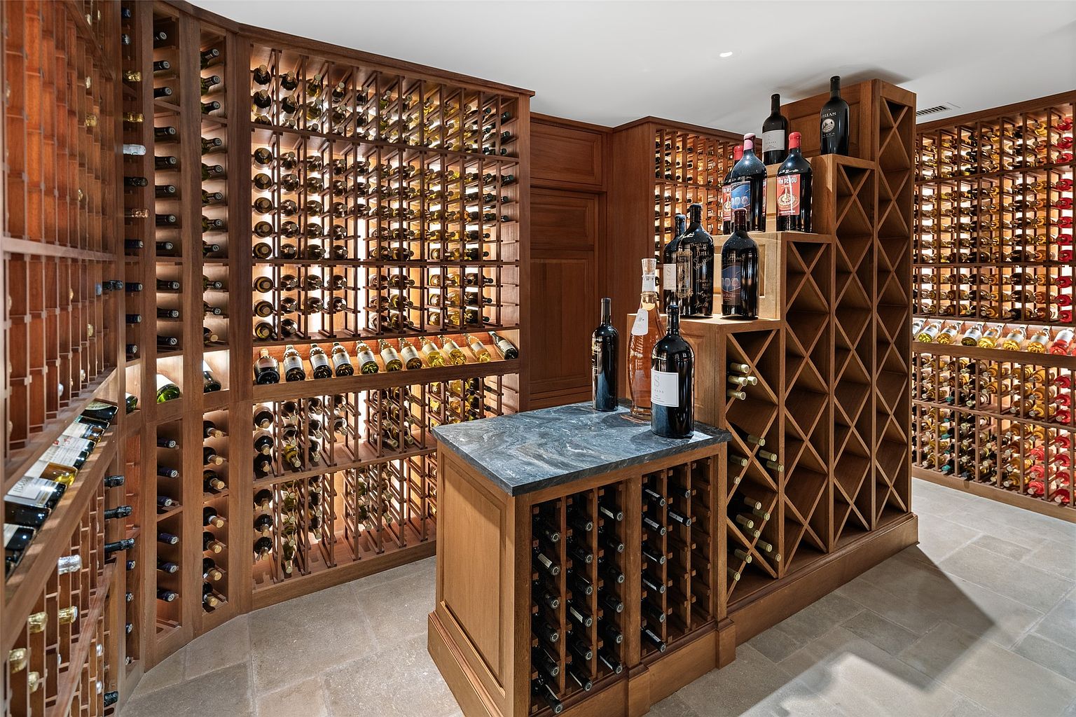 This is a well-organized wine cellar featuring custom wooden racks filled with numerous wine bottles. The cellar includes a central island with a dark countertop, providing a space for tasting or display. The warm lighting and stone flooring create an inviting and sophisticated atmosphere, perfect for wine enthusiasts.
