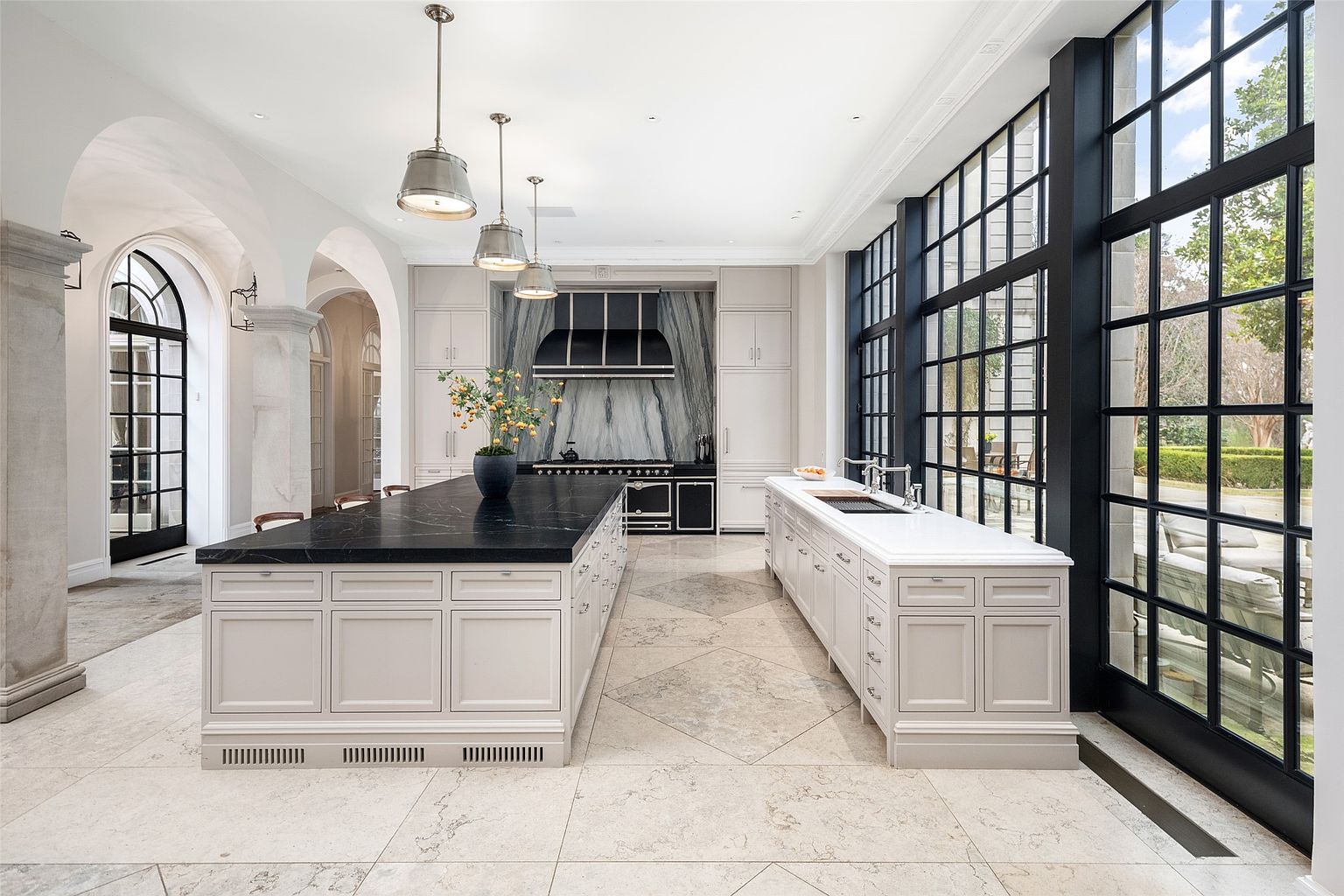 This is a bright and spacious kitchen featuring a large island with a dark countertop and custom cabinetry. The kitchen also includes a long counter with a sink, a black range, and a wall of windows that provide natural light and views of the outdoors. The flooring is a light-colored tile, and the overall style is luxurious and elegant.