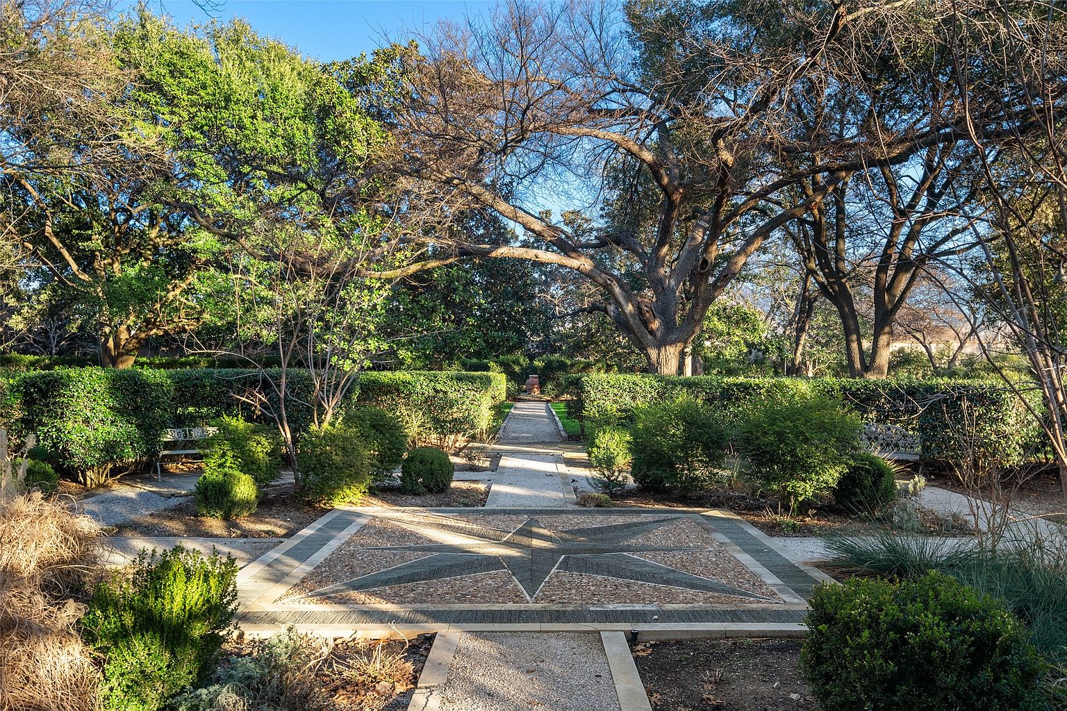 This image showcases a meticulously designed garden with a symmetrical layout. A central pathway, adorned with a star-patterned stone mosaic, leads through manicured hedges and lush greenery. Mature trees provide shade and create a serene atmosphere, enhancing the property's appeal.