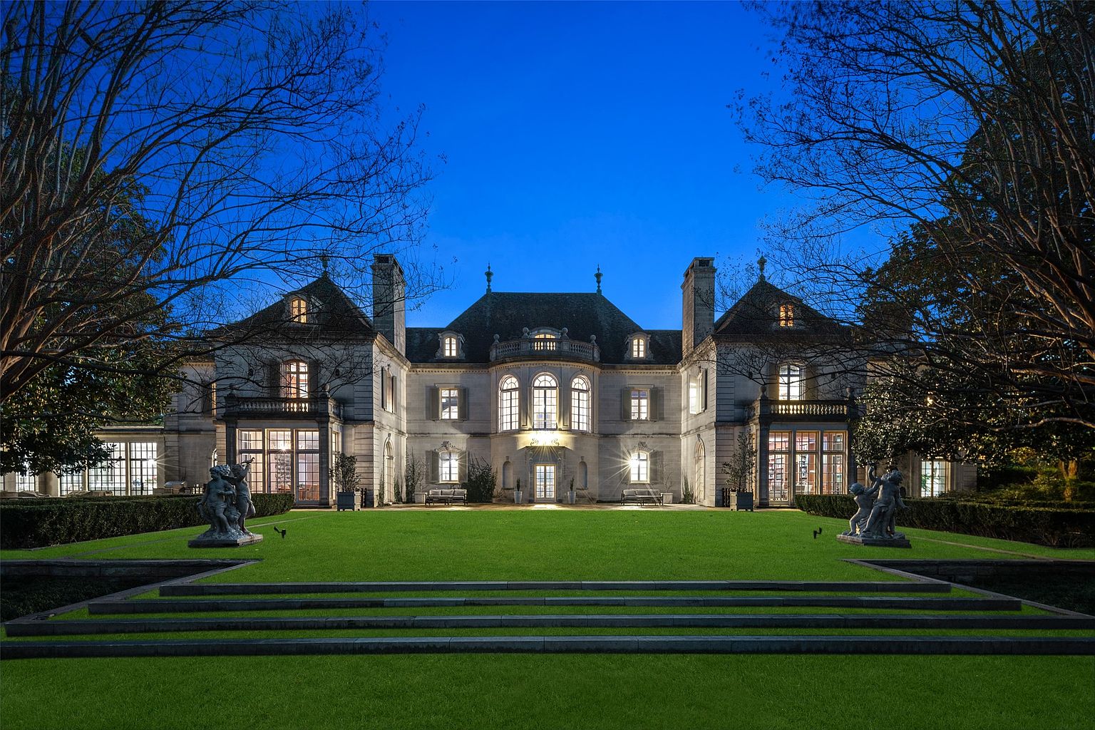 This is a grand, symmetrical front view of a large estate home at dusk. The architecture features a stone facade, multiple windows with lights on, and a manicured lawn leading up to the house. Statues flank the lawn, and the overall impression is one of luxury and elegance.