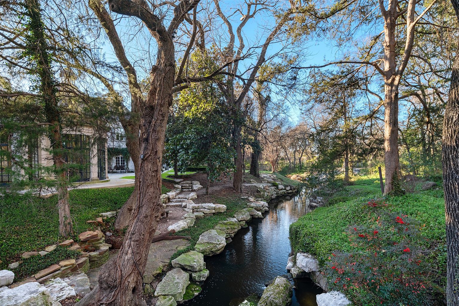 This image showcases a serene garden setting with a flowing stream, lush greenery, and mature trees. Stone pathways and rock formations add to the natural aesthetic, creating a tranquil and inviting outdoor space. A glimpse of the house in the background suggests a seamless integration of the property with its natural surroundings.