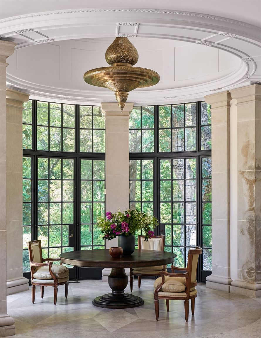 This interior shot showcases an elegant dining room with a large round table centered beneath an ornate chandelier. The room is surrounded by tall windows offering a view of lush greenery, complemented by classic chairs and architectural columns. The space exudes a sense of refined luxury and tranquility, ideal for sophisticated gatherings.