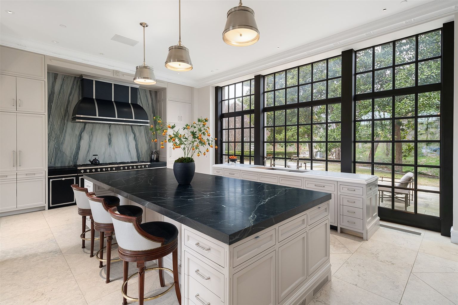 This is a bright and spacious kitchen featuring a large island with a dark countertop and seating. The kitchen is equipped with high-end appliances and custom cabinetry, complemented by a striking marble backsplash and a large window offering natural light and views of the outdoors. The overall design exudes luxury and sophistication, making it a desirable space for cooking and entertaining.