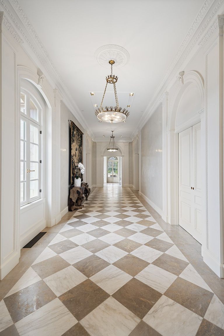 This is a grand hallway featuring a checkerboard marble floor, ornate ceiling moldings, and elegant lighting fixtures. The walls are painted in a neutral tone, and arched doorways add to the architectural detail. The hallway appears spacious and luxurious, creating a sophisticated and inviting atmosphere.