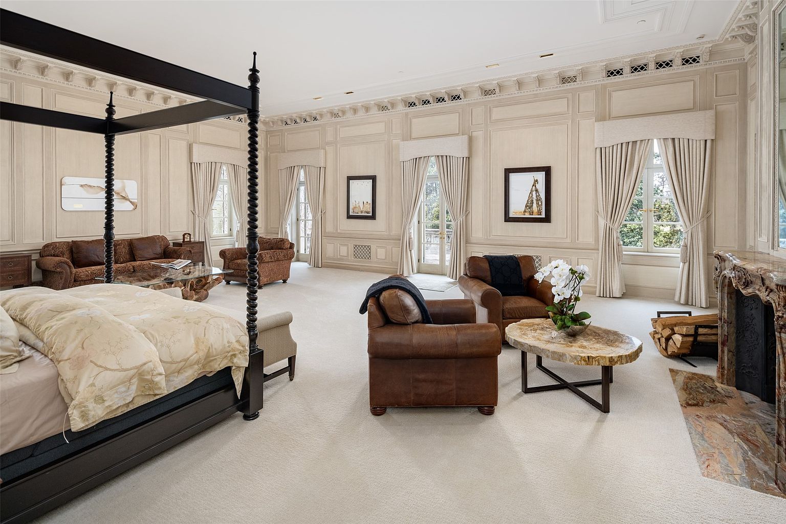 This is a spacious primary bedroom featuring a four-poster bed with luxurious bedding, complemented by a seating area with plush armchairs and a sofa. The room is decorated in neutral tones with detailed wall paneling and crown molding, creating an elegant and comfortable atmosphere. A fireplace adds a touch of warmth and sophistication to the space.