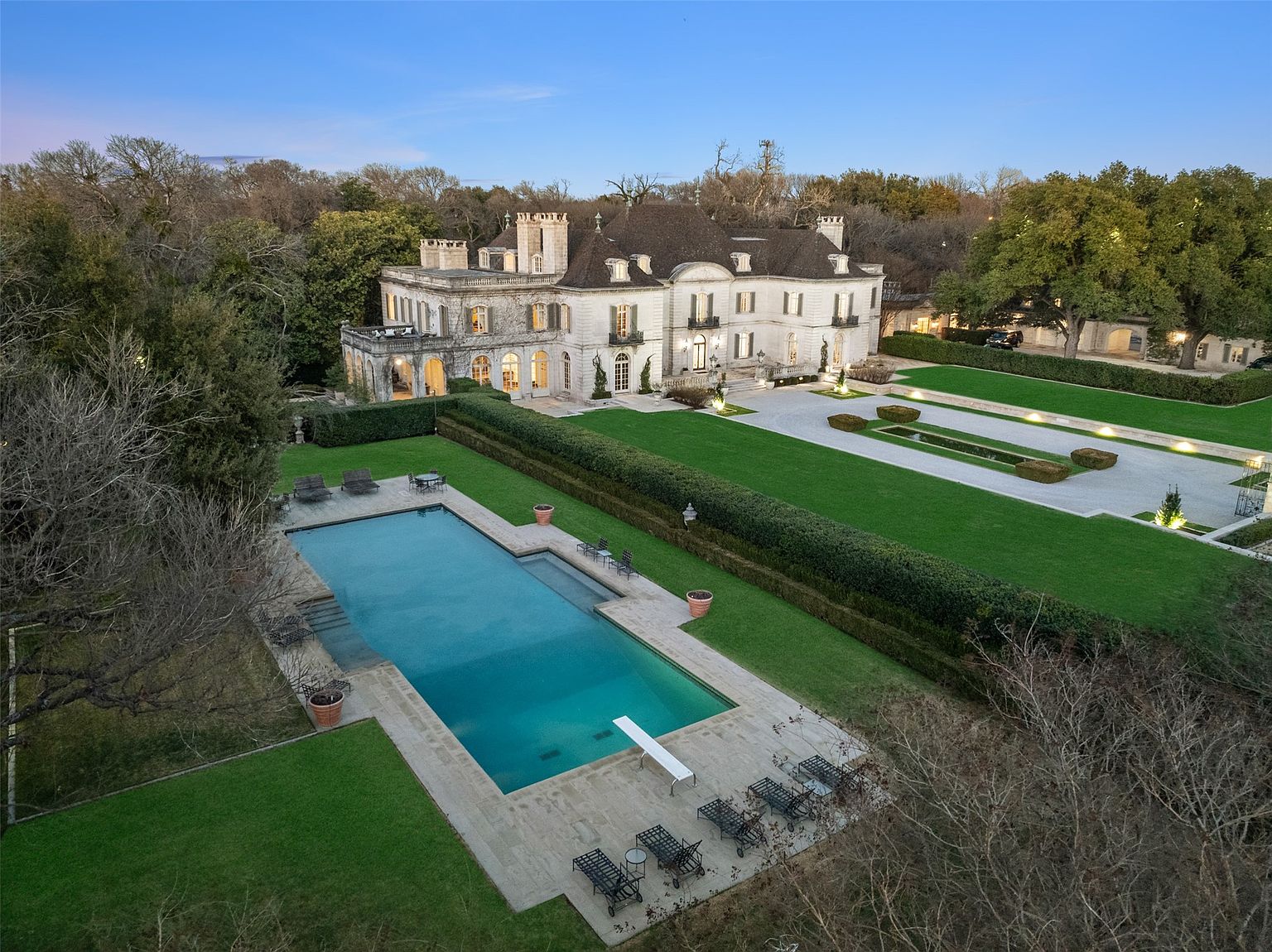 This aerial view showcases a grand estate with a large swimming pool, meticulously manicured lawns, and formal gardens. The stately home features a light-colored facade, multiple stories, and intricate architectural details. The property is surrounded by mature trees, providing privacy and a sense of seclusion.