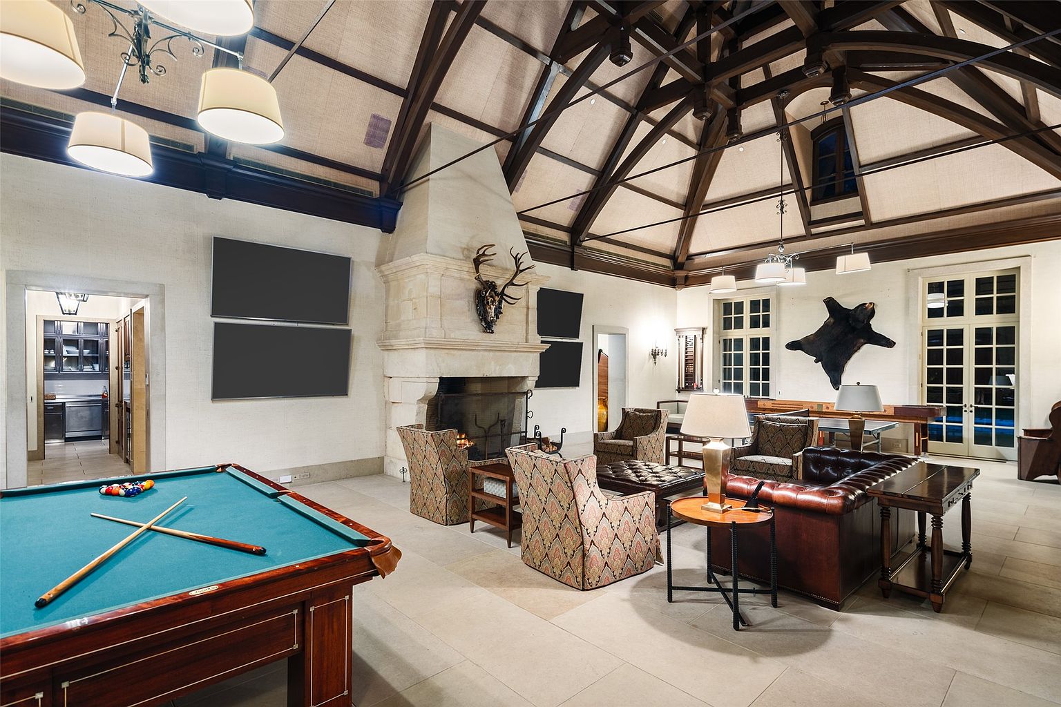 This grand living room features a high vaulted ceiling with exposed wooden beams, a stone fireplace with mounted antlers, and multiple seating areas. A pool table adds a recreational element, while large windows provide ample natural light. The room exudes a rustic yet luxurious atmosphere, perfect for entertaining or relaxing.