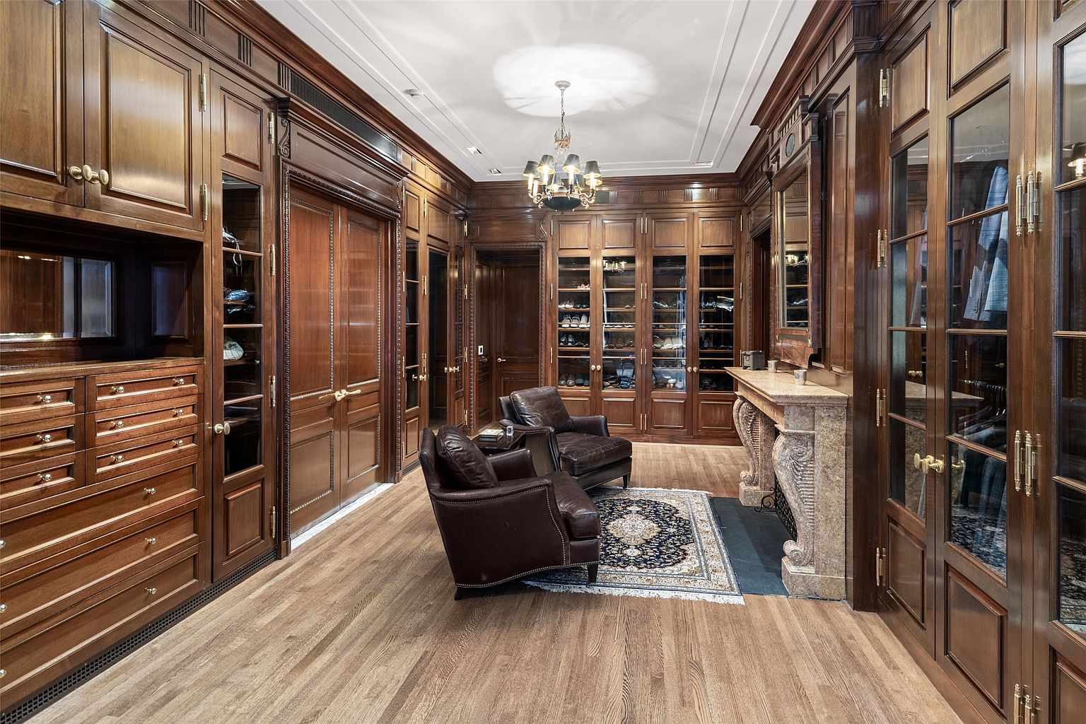 This is a luxurious walk-in closet featuring custom wood cabinetry with glass doors and drawers. Two leather armchairs sit on a patterned rug in the center of the room, adding a touch of elegance. A decorative fireplace mantel is visible, and the hardwood flooring adds warmth to the space.