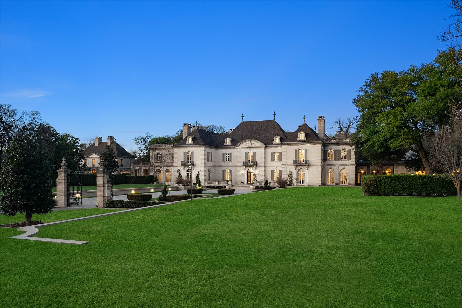 This image showcases the grand front exterior of a large estate home. The architecture features a symmetrical design with multiple windows, balconies, and a prominent roofline. A sprawling green lawn leads up to the house, complemented by manicured landscaping and a gated entrance, creating an impression of luxury and privacy.