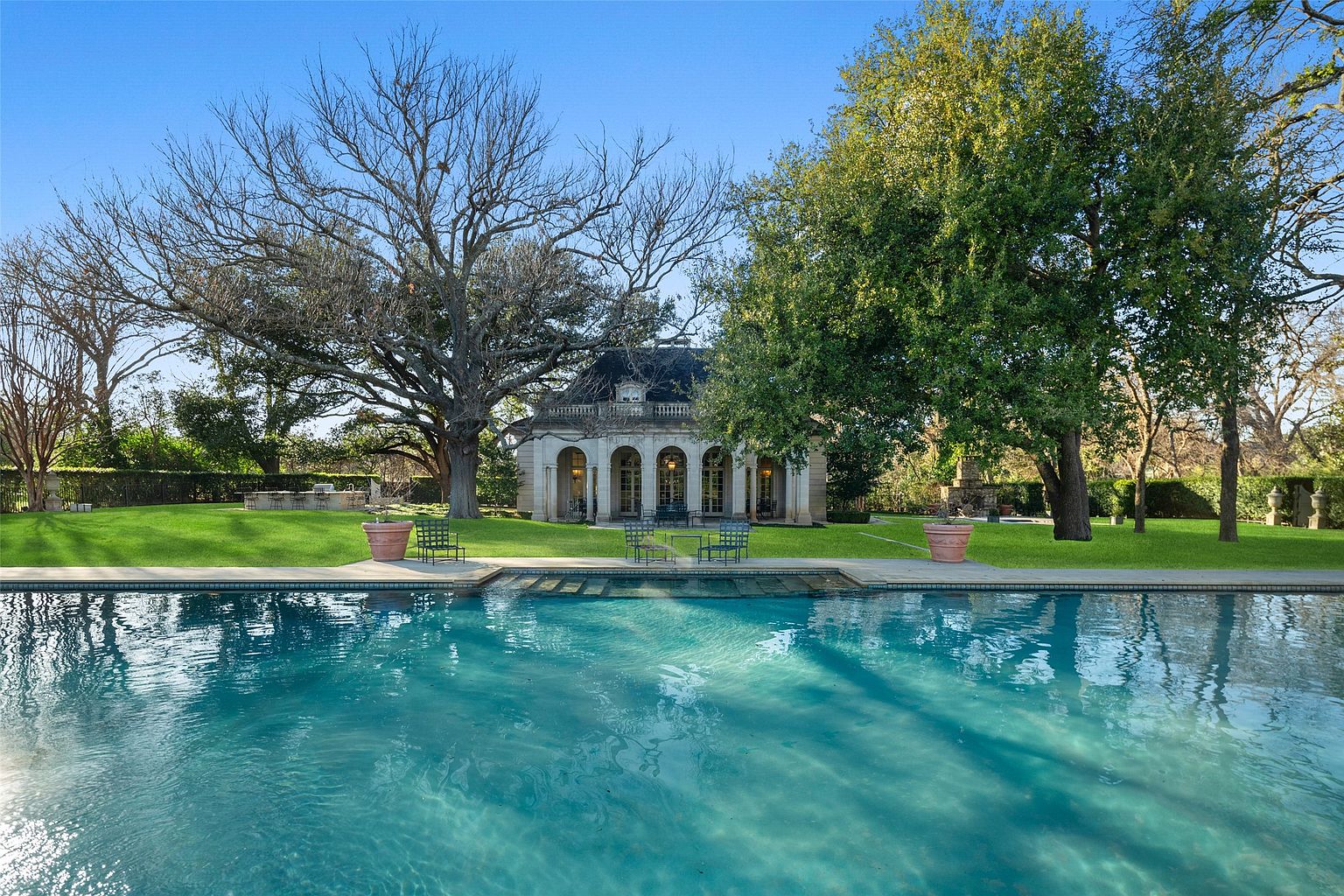 This image showcases a luxurious pool area with a classical-style pool house in the background. The pool's turquoise water reflects the sky, creating a serene atmosphere. Lush green lawns surround the pool, and mature trees provide shade and privacy, enhancing the property's appeal.