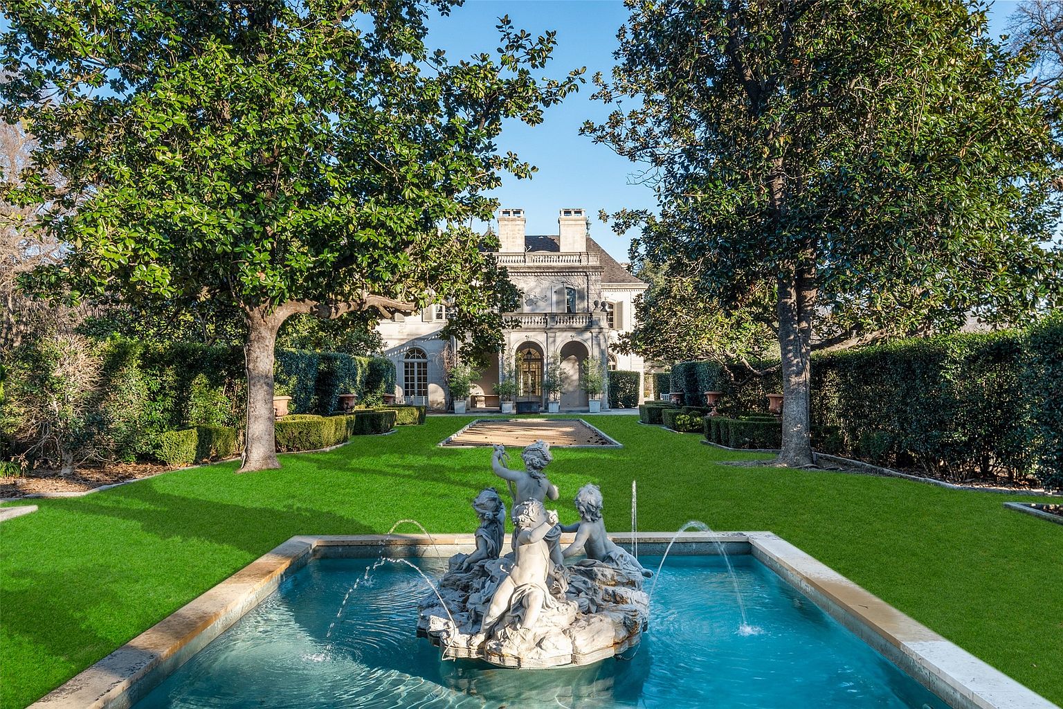 This exterior shot showcases a meticulously landscaped yard with a grand fountain as the focal point. Lush green lawns are bordered by manicured hedges and mature trees, leading to an elegant home in the background. The scene exudes a sense of luxury and tranquility, highlighting the property's outdoor living space.