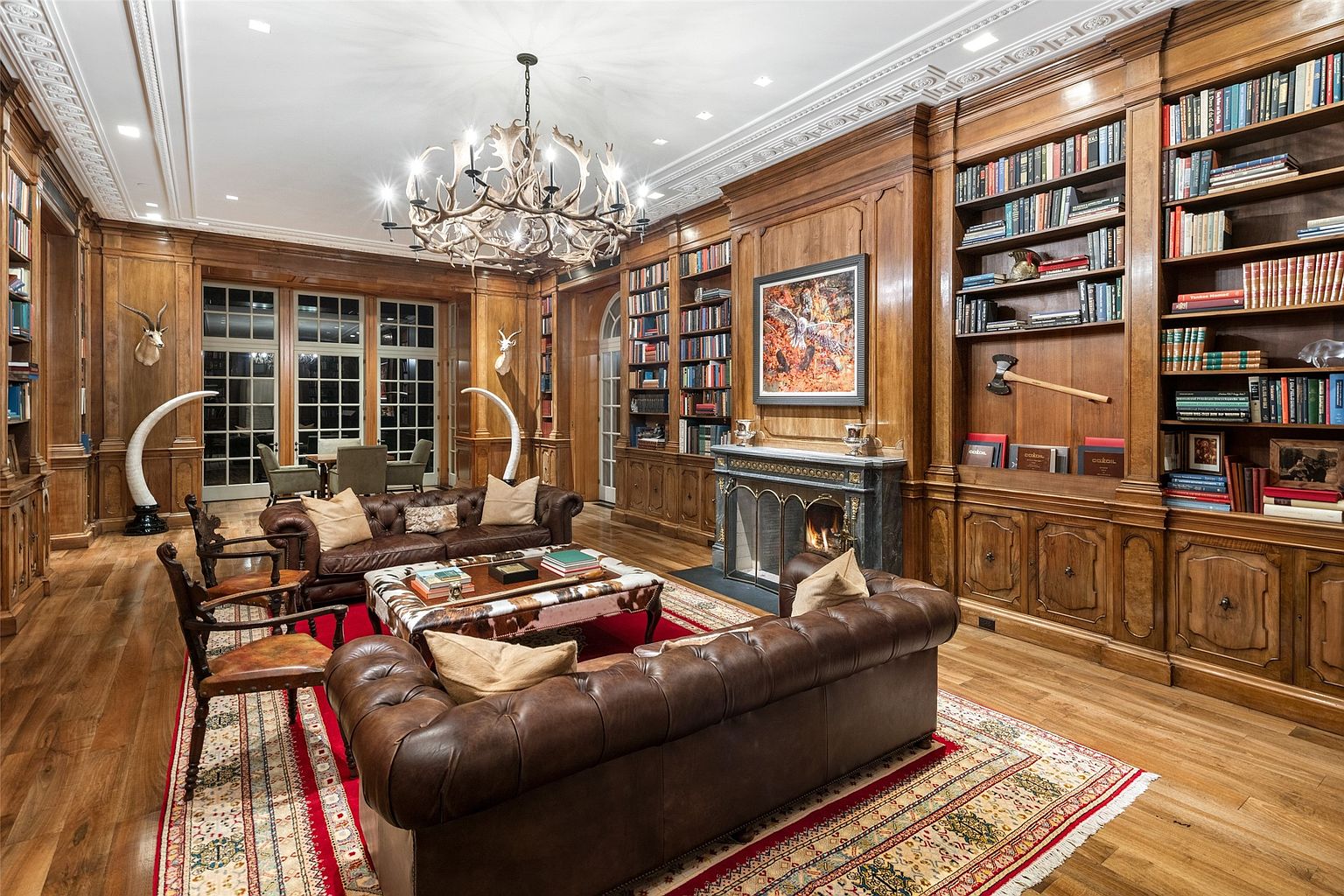 This is a luxurious living room featuring wood-paneled walls, built-in bookshelves, and a grand antler chandelier. Two leather sofas and a patterned ottoman create a comfortable seating area around a fireplace. The room exudes a classic, sophisticated style with a touch of rustic charm.