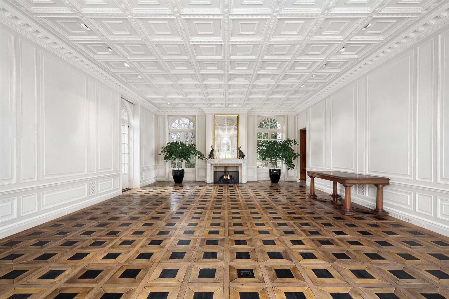 This grand hallway features a striking geometric patterned wood floor and ornate white wall paneling. The coffered ceiling adds a touch of elegance, while the fireplace and arched windows create a focal point. A wooden table sits to the right, adding a touch of warmth to the otherwise bright and airy space.