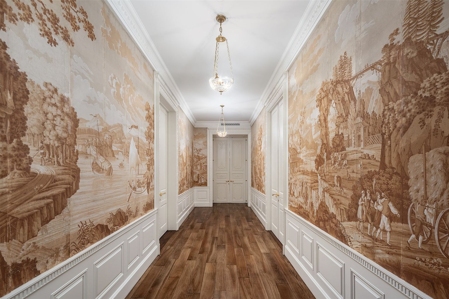 This is a beautifully decorated hallway featuring scenic wallpaper, white wainscoting, and hardwood floors. The hallway is illuminated by elegant pendant lighting, creating a warm and inviting atmosphere. Multiple doorways line the hallway, suggesting access to various rooms within the home.