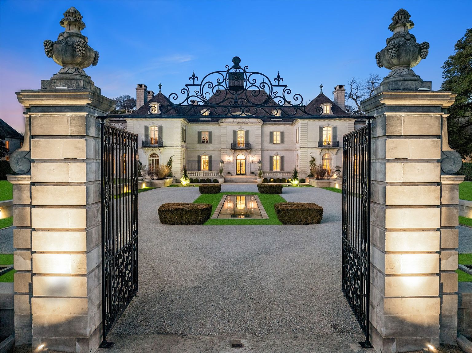 This is a grand front view of a luxurious estate, showcasing an elegant symmetrical facade with multiple windows and balconies. The property is accessed through ornate wrought iron gates flanked by substantial stone pillars topped with decorative urns. A meticulously landscaped driveway and garden with a reflecting pool lead to the impressive residence, creating a striking first impression.