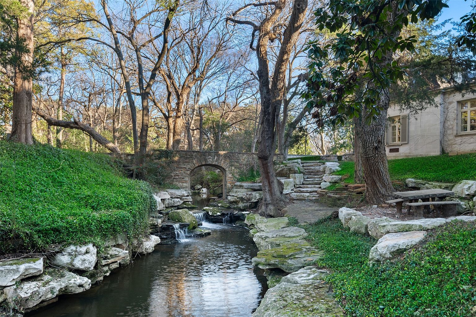 This picturesque yard/garden features a stone bridge over a gently flowing stream with small waterfalls, surrounded by lush greenery and mature trees. A stone staircase leads up to a building, and a stone bench offers a place to relax and enjoy the serene setting. The scene evokes a sense of tranquility and natural beauty, making it an appealing outdoor space.