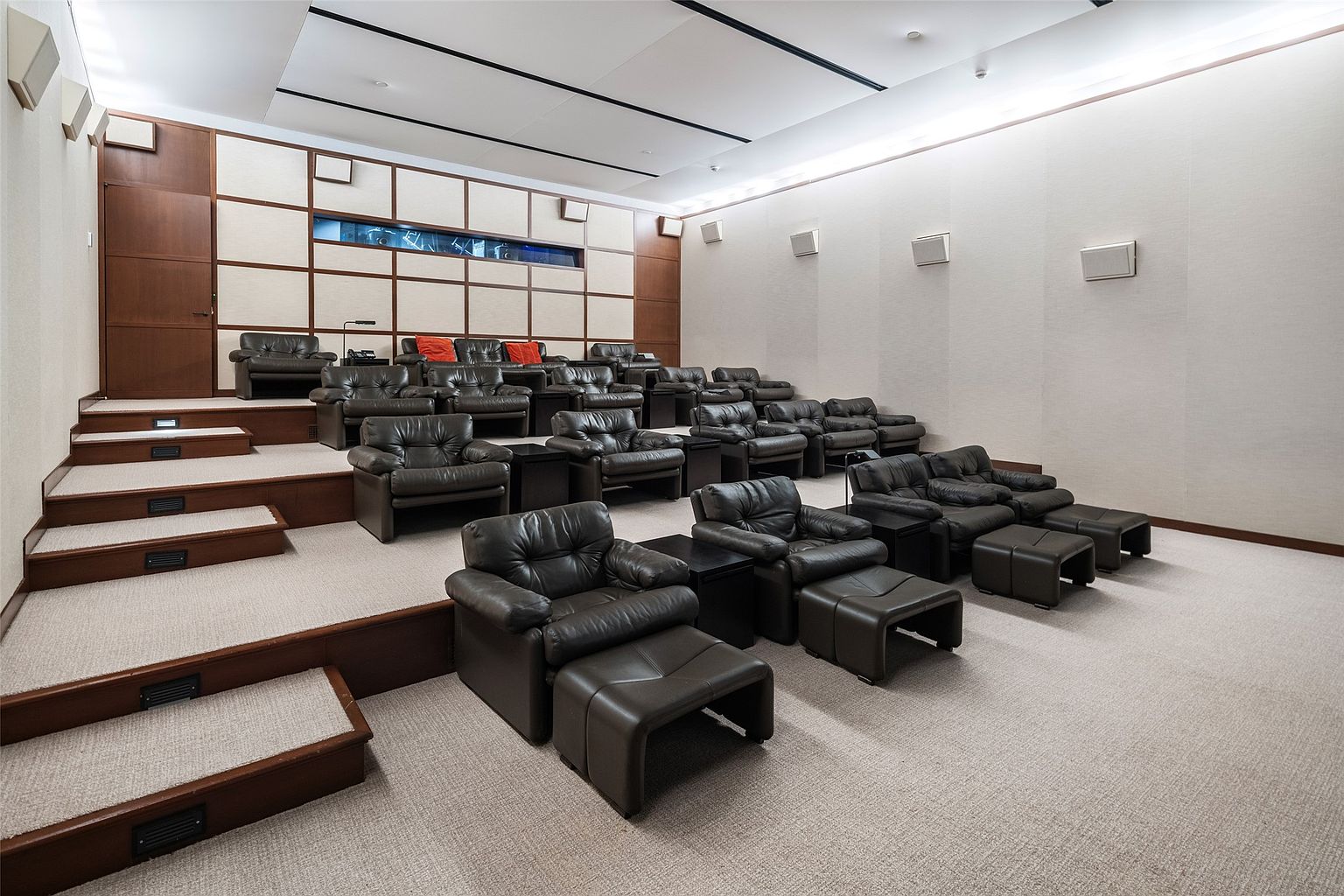 This is an interior shot of a home theater, featuring tiered seating with plush, dark leather armchairs and ottomans. The room is designed for optimal viewing, with acoustic paneling on the walls and a neutral color palette. The overall impression is one of luxury and comfort, perfect for a cinematic experience.