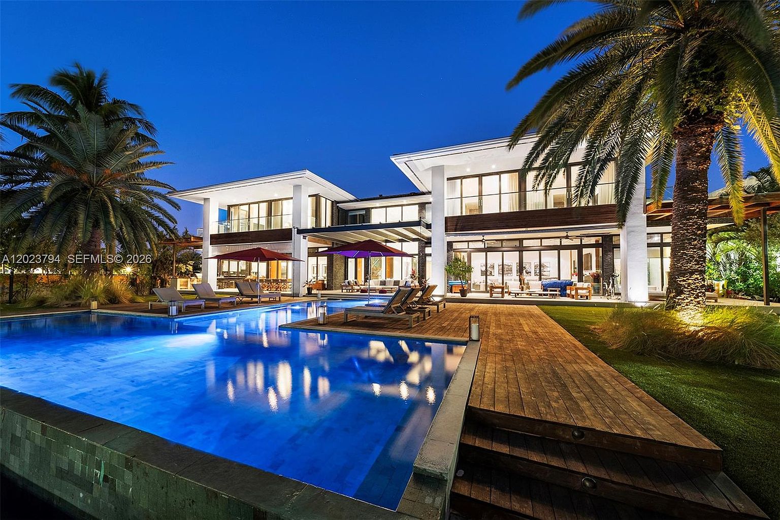 This stunning contemporary residence is captured at twilight, showcasing a sprawling multi-level home with expansive glass walls and a large, illuminated infinity-edge pool. The foreground features a rich wood deck with lounge seating, while lush palm trees and manicured landscaping frame the architectural structure. The scene exudes a luxurious, resort-like atmosphere, emphasizing the seamless transition between indoor and outdoor living spaces.