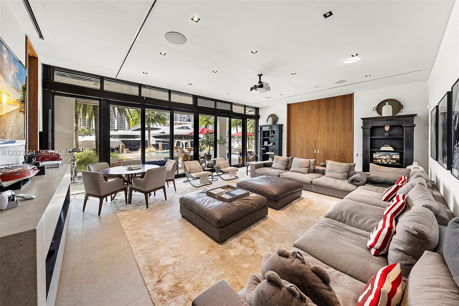 This expansive, open-concept living room features a large, neutral-toned sectional sofa adorned with red-striped accent pillows and two matching leather ottomans. The space is illuminated by recessed lighting and a projector mounted on the ceiling, with floor-to-ceiling glass doors providing a seamless transition to the outdoor area. A dining table sits near the windows, while a dark, traditional-style shelving unit with a built-in television adds a classic contrast to the modern, light-filled interior.