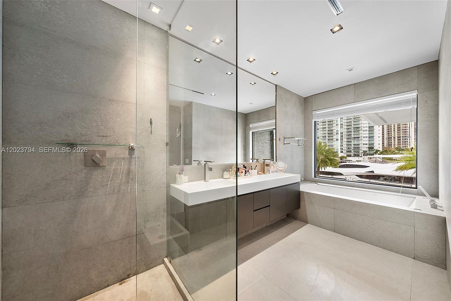 This modern primary bathroom features a sleek, floating vanity with a double sink and a large, frameless glass-enclosed shower. A deep soaking tub is positioned beneath a large window that offers a scenic view of the marina and surrounding buildings. The space is finished with neutral-toned, large-format wall and floor tiles, creating a clean and sophisticated aesthetic.
