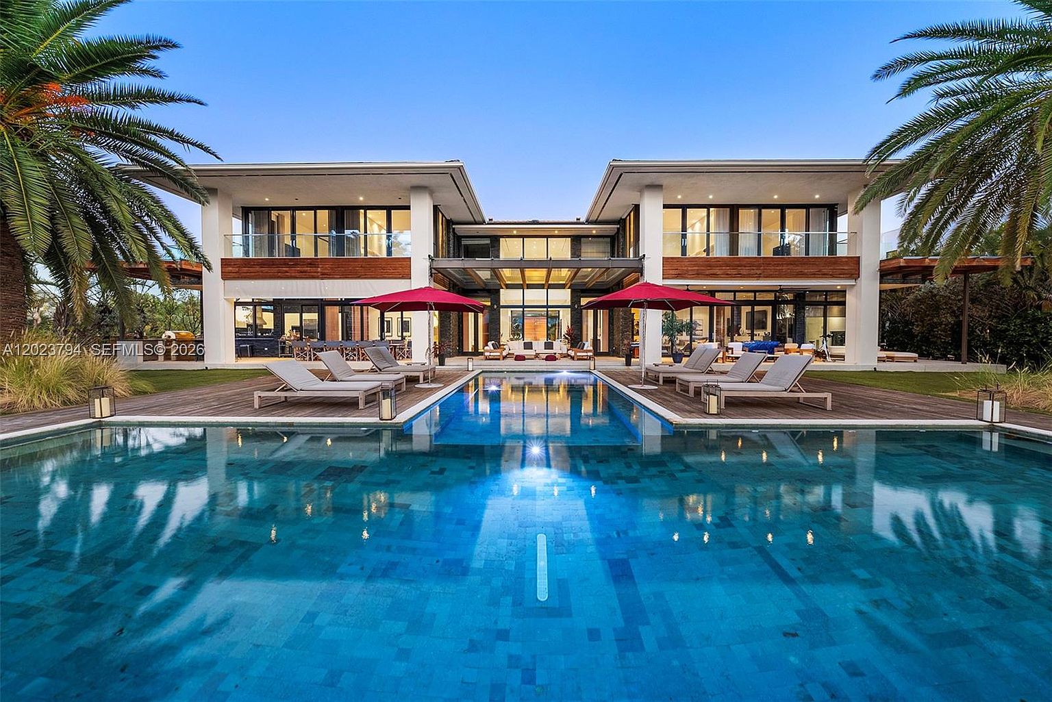 This stunning modern estate features a symmetrical architectural design with a large, rectangular swimming pool as the central focal point. The rear facade showcases extensive use of glass, including large windows and sliding doors that open onto a spacious wooden deck furnished with lounge chairs and red umbrellas. Lush palm trees frame the property, creating a luxurious and serene atmosphere at dusk.