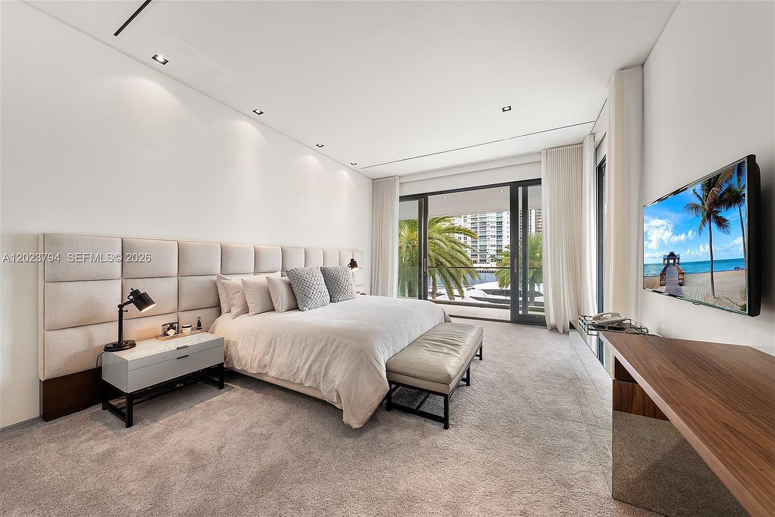 This spacious primary bedroom features a large, upholstered headboard wall and a neutral color palette that creates a serene atmosphere. A set of floor-to-ceiling glass doors opens onto a balcony, providing a view of the water and palm trees outside. The room is furnished with a modern bed, a sleek bedside table with a lamp, and a wooden desk area, all complemented by soft carpeting.