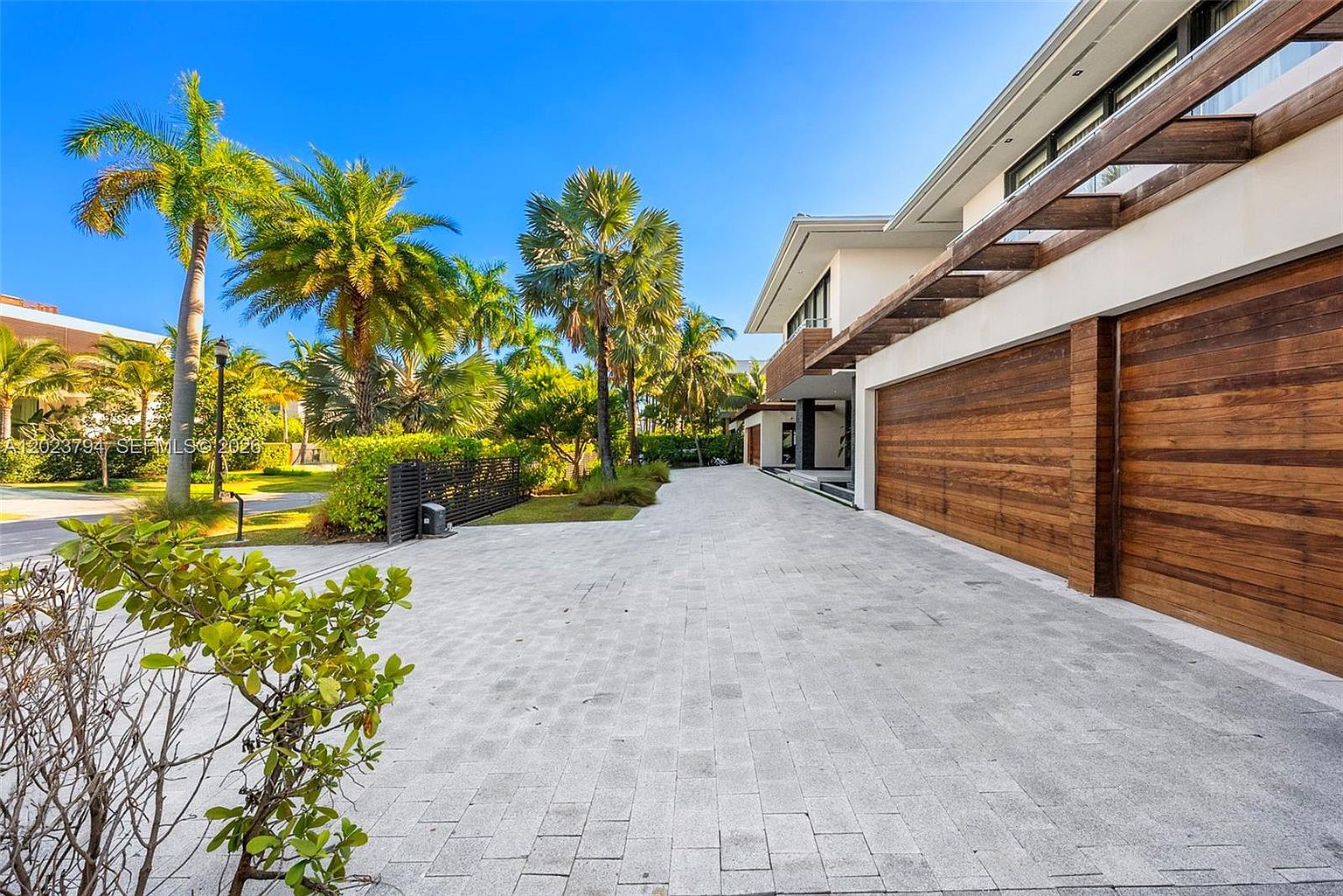 This exterior view captures a modern home's expansive driveway leading to a multi-car garage with warm-toned wood paneling. The architecture features clean lines and a prominent wooden pergola structure extending over the garage doors. Lush tropical landscaping, including tall palm trees, frames the paved driveway, creating a bright and inviting atmosphere under a clear blue sky.