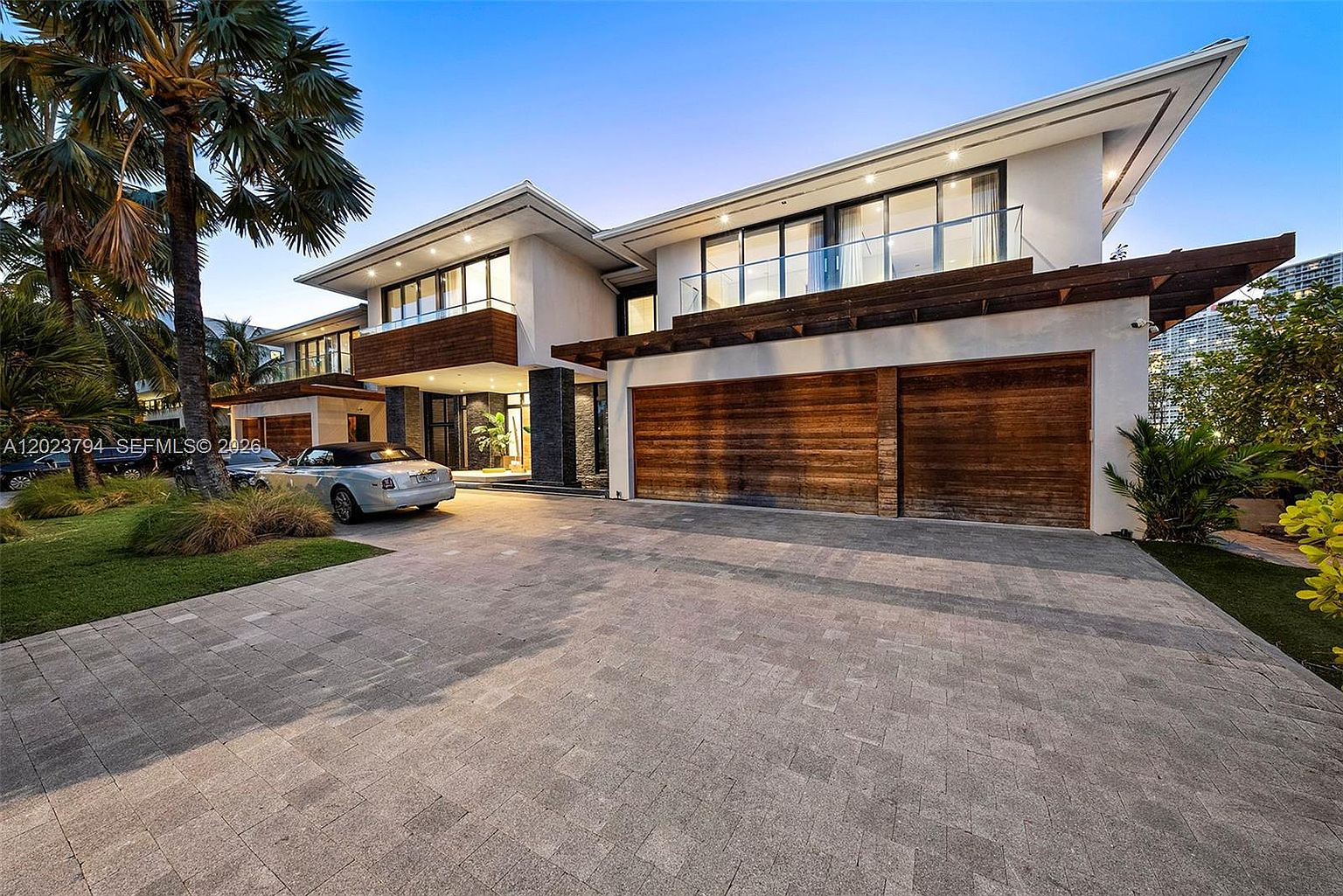 This modern luxury residence features a striking architectural design with clean lines, a combination of smooth white stucco, and warm wood-paneled garage doors. The front facade includes a spacious paved driveway, a prominent palm tree, and large windows that reflect the evening sky. The overall impression is one of contemporary elegance and high-end curb appeal.
