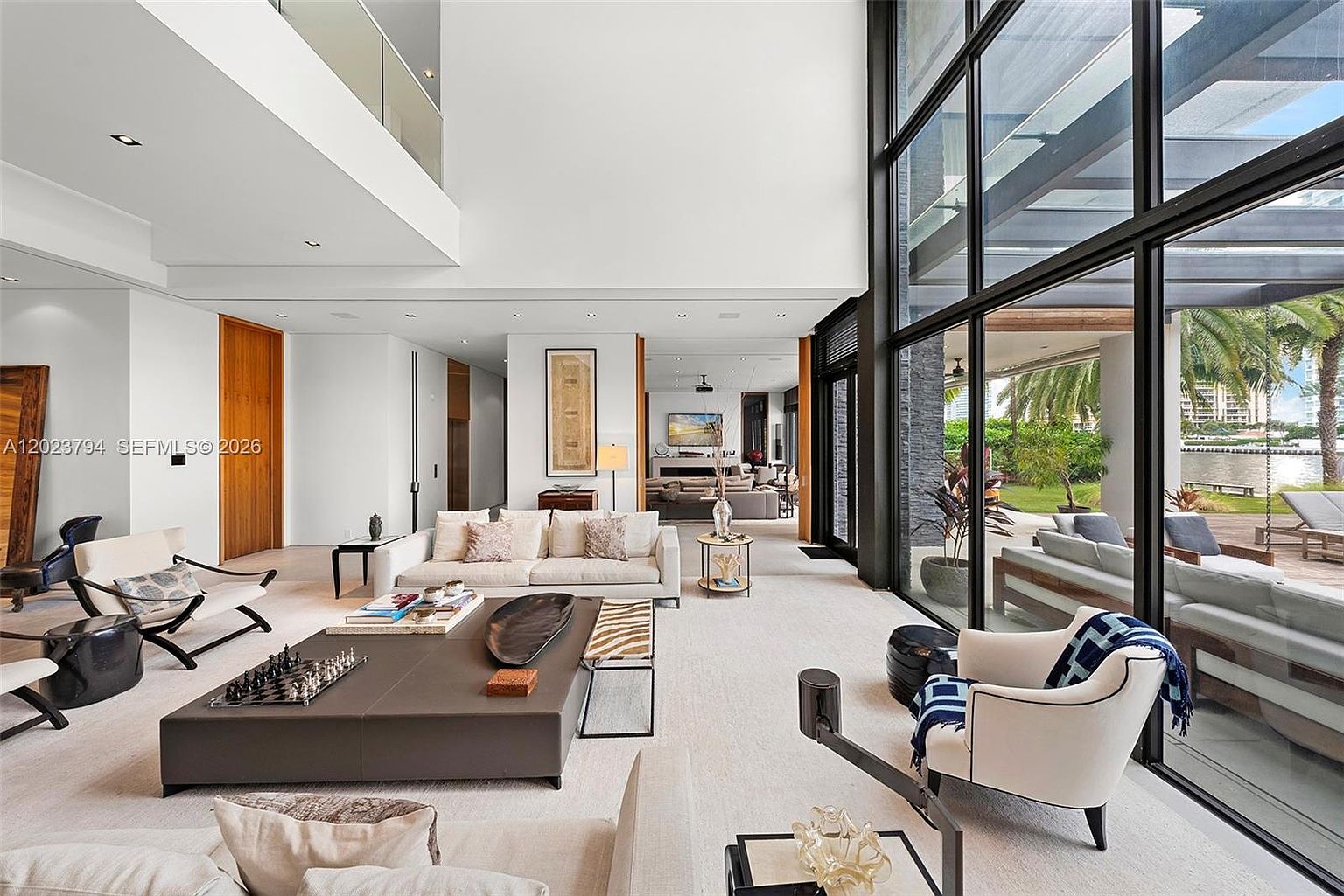 This expansive, double-height living room features a minimalist aesthetic with neutral tones and a large, low-profile sectional sofa. A wall of floor-to-ceiling glass panels provides an unobstructed view of the outdoor patio and waterfront beyond, flooding the space with natural light. The room is anchored by a large, dark-toned coffee table and a white armchair, creating a sophisticated and airy atmosphere.