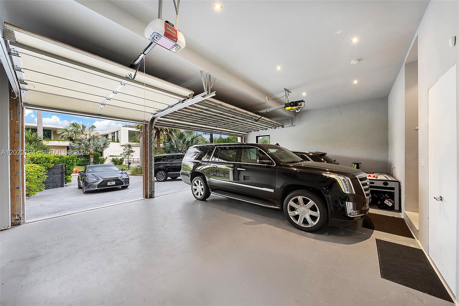 This spacious, well-lit garage features a smooth, neutral-toned floor and a high ceiling equipped with modern lighting and overhead door mechanisms. A large black SUV is parked inside, while the open garage door reveals a paved driveway with another vehicle parked outside amidst lush tropical landscaping. The perspective is from inside the garage looking out, highlighting the seamless transition between the indoor storage space and the exterior environment.