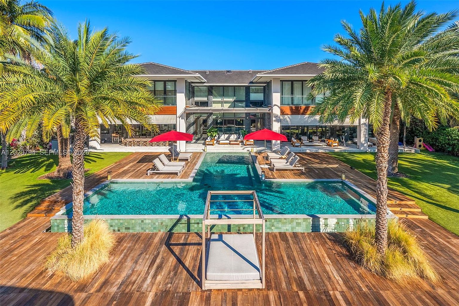 This expansive rear exterior view showcases a modern luxury residence featuring a large, geometric swimming pool as the centerpiece. The pool is surrounded by a spacious wooden deck with multiple lounge chairs, red umbrellas, and a central cabana, all set against a backdrop of lush green lawn and tall palm trees. The home itself displays a contemporary architectural style with large glass panels and clean lines, creating a seamless transition between indoor and outdoor living spaces.