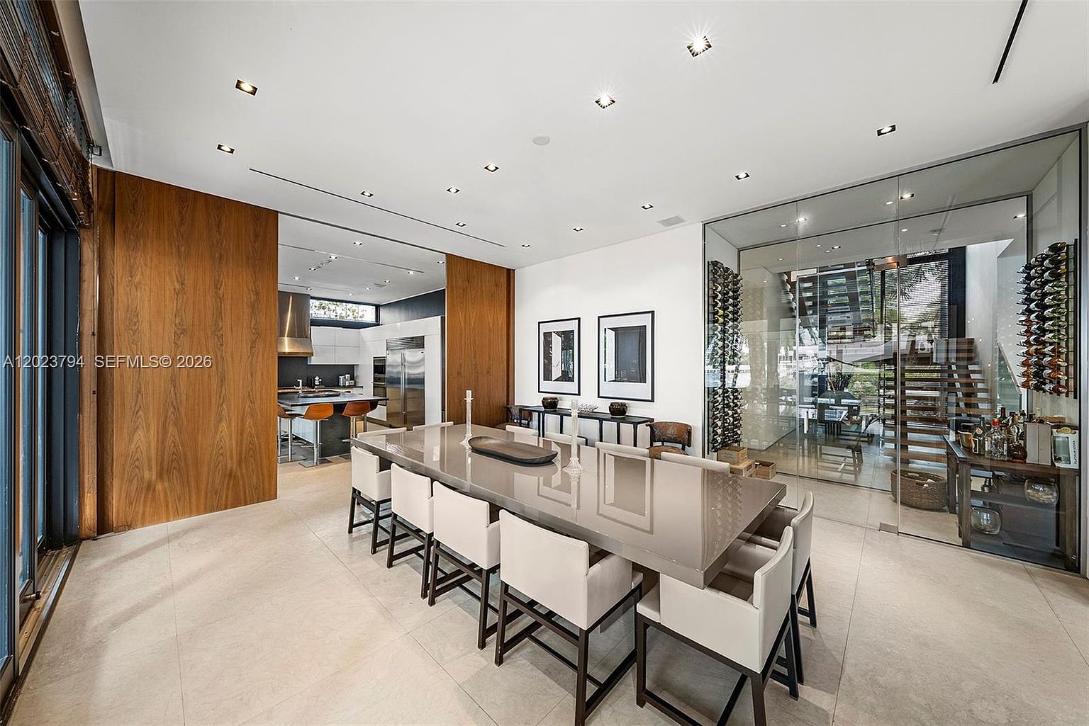 This sophisticated dining area features a long, glossy table surrounded by eight modern chairs with light-colored upholstery and dark frames. To the right, a glass-enclosed wine cellar adds a dramatic architectural element, while the left side opens into a sleek kitchen with wood-paneled walls. The space is illuminated by recessed ceiling lights, creating a bright and polished atmosphere suitable for formal entertaining.