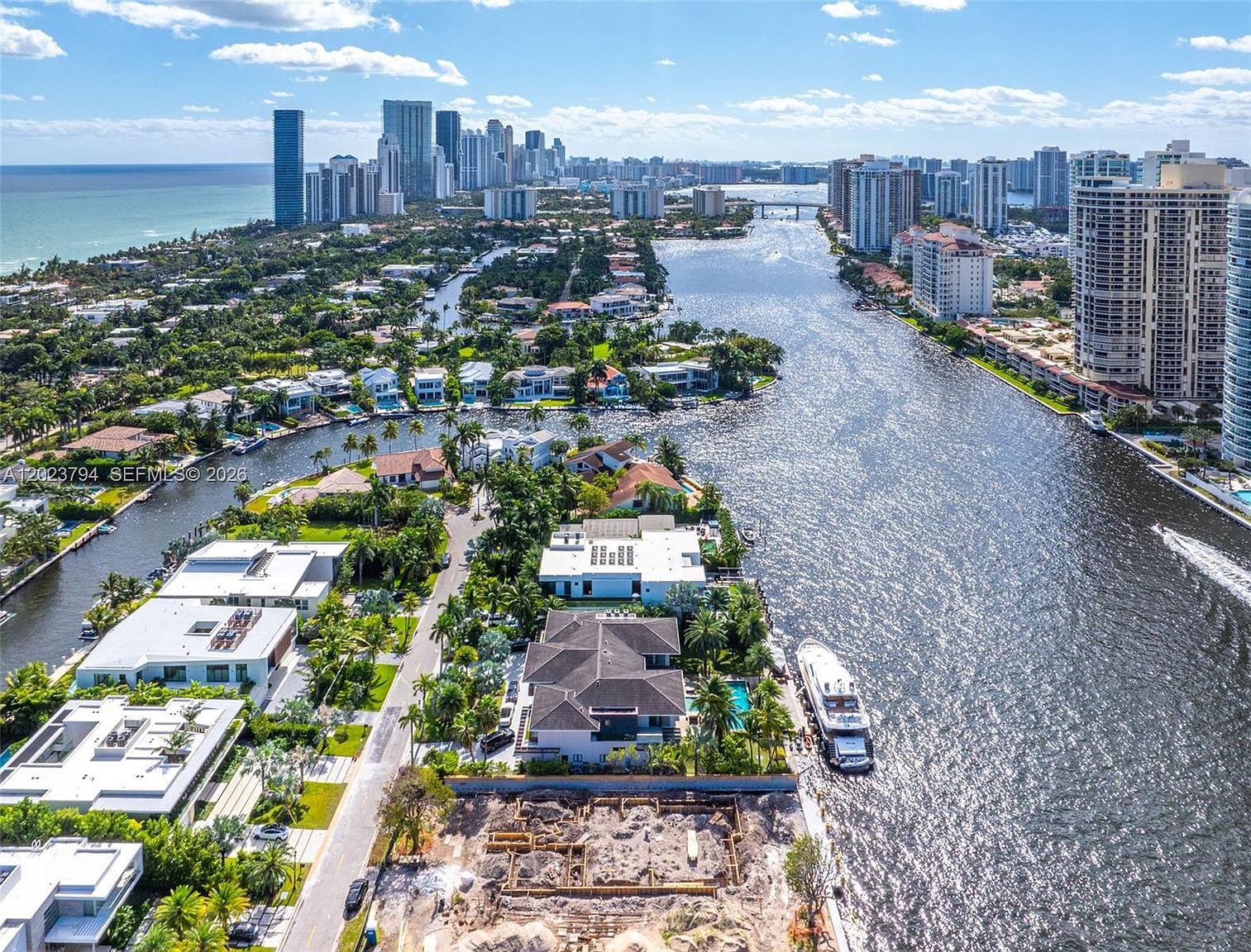This high-altitude aerial view captures a luxurious waterfront neighborhood in a coastal city, showcasing a mix of modern estates and high-rise condominiums. The scene features a sparkling waterway filled with boats, lush tropical landscaping, and a prominent construction site in the foreground. The perspective emphasizes the prime location and the expansive, sun-drenched urban landscape.