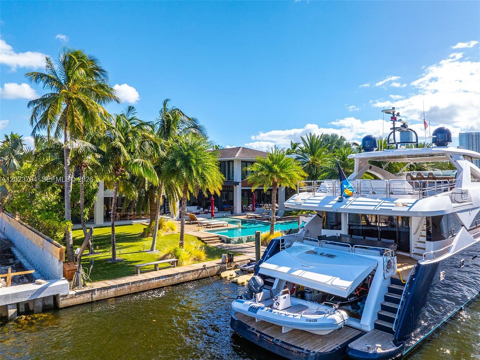 This high-angle, cinematic shot captures a luxurious waterfront property featuring a large, modern residence with a private dock and a swimming pool. A substantial yacht is moored directly alongside the property's bulkhead, emphasizing the exclusive lifestyle and deep-water access. The lush, manicured lawn is dotted with mature palm trees, creating a tropical resort-like atmosphere that seamlessly blends the indoor living spaces with the outdoor pool deck.
