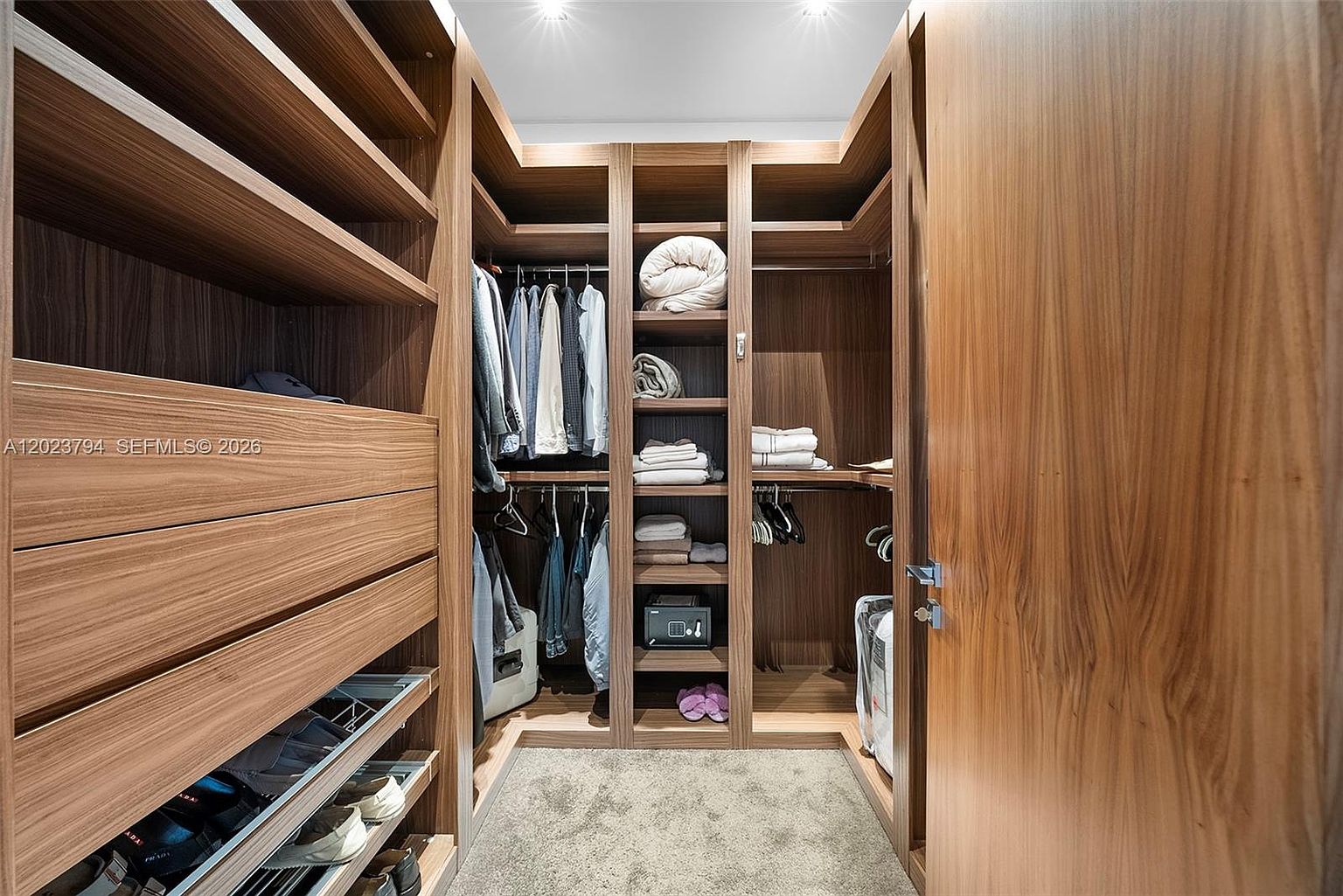 This walk-in closet features custom wood cabinetry with a mix of hanging rods, open shelving, and pull-out drawers. The space is organized with folded linens, hanging garments, and a small safe, all set against a neutral carpeted floor. The warm wood tones and recessed lighting create a sophisticated and highly functional storage environment.