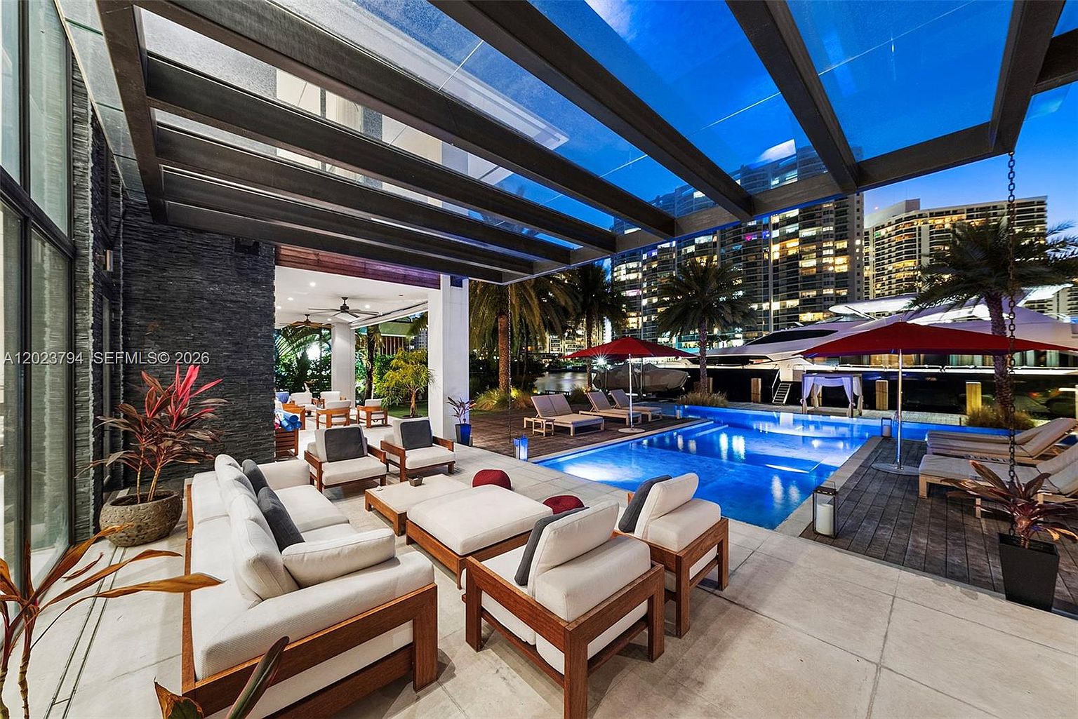 This luxurious outdoor living space features a covered patio area with comfortable lounge seating, seamlessly transitioning to a private swimming pool overlooking a waterfront marina. The area is defined by a modern pergola structure, dark stone wall accents, and a wooden deck, creating a sophisticated atmosphere for relaxation. The perspective captures the inviting lounge furniture in the foreground, leading the eye toward the illuminated pool and the docked yachts in the background.
