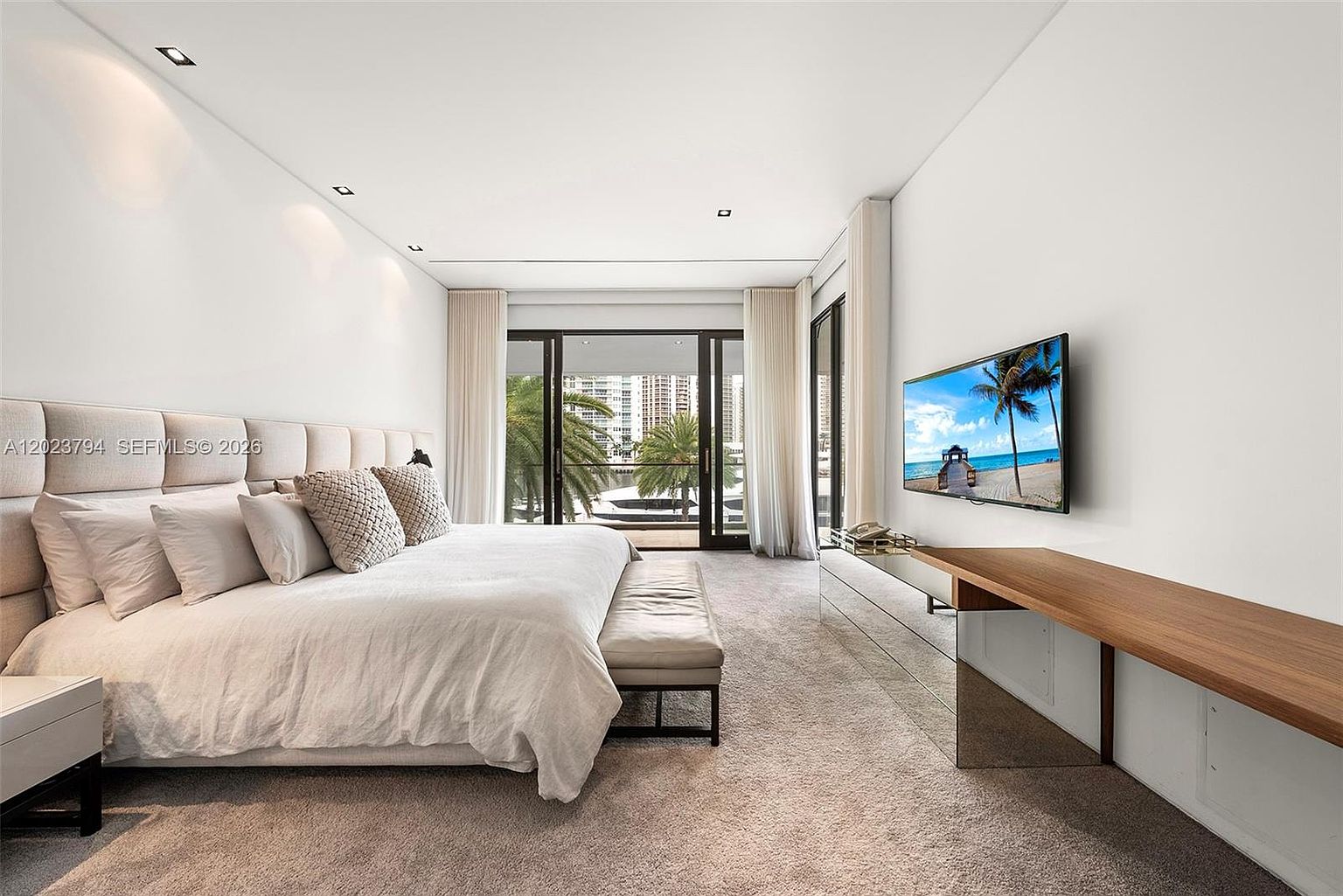 This spacious primary bedroom features a neutral color palette with a large, upholstered headboard spanning the width of the wall. A sleek, low-profile wooden console sits beneath a wall-mounted television, while floor-to-ceiling glass doors provide access to a private balcony with tropical views. The room is finished with plush carpeting and soft, layered window treatments, creating a serene and sophisticated atmosphere.