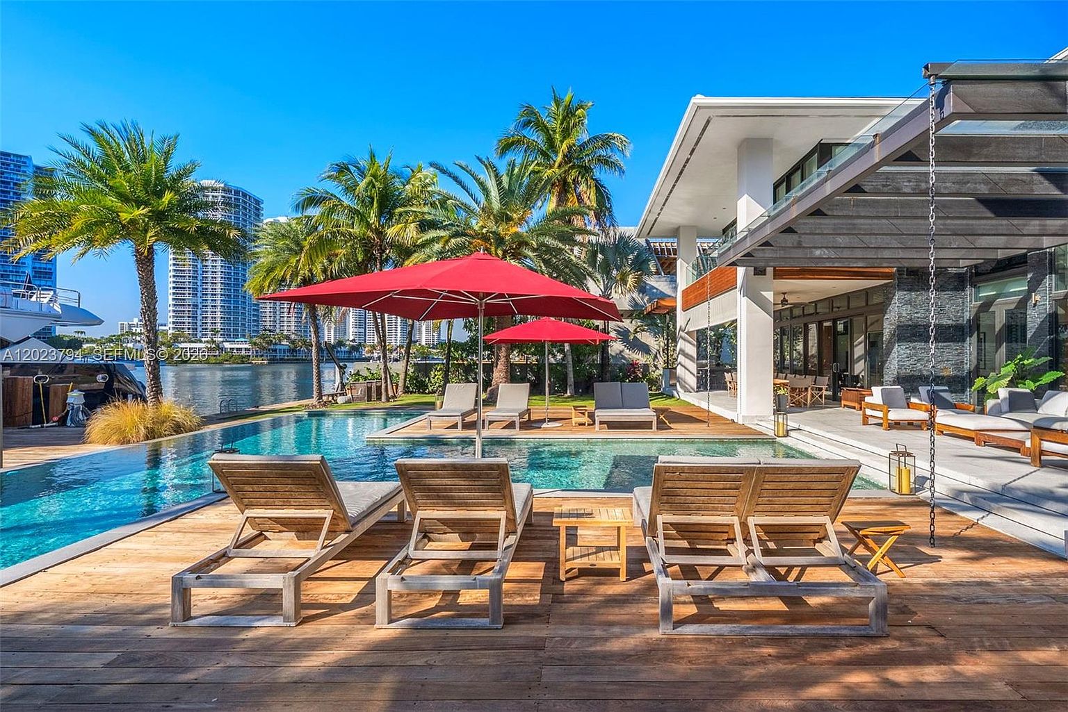 This luxurious waterfront outdoor living space features a sleek, rectangular swimming pool set into a spacious wooden deck. The area is furnished with multiple lounge chairs and vibrant red umbrellas, creating a resort-like atmosphere overlooking a canal and city skyline. The modern architecture of the home includes a covered patio area with an open-concept design, seamlessly blending indoor and outdoor living.