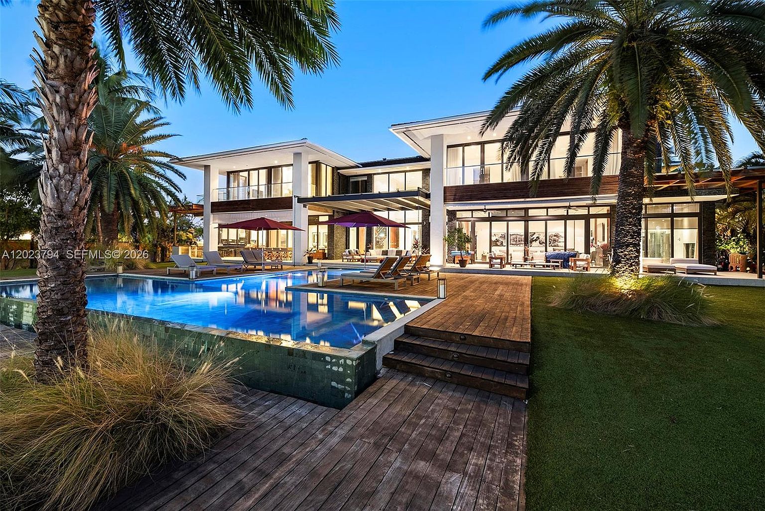 This stunning contemporary estate features a sprawling rear exterior with a large, rectangular swimming pool as the focal point. The home showcases modern architectural lines, extensive glass walls, and a multi-level design with wooden decking and manicured landscaping. The scene is captured at twilight, highlighting the warm interior lighting against the cool blue tones of the pool and evening sky.